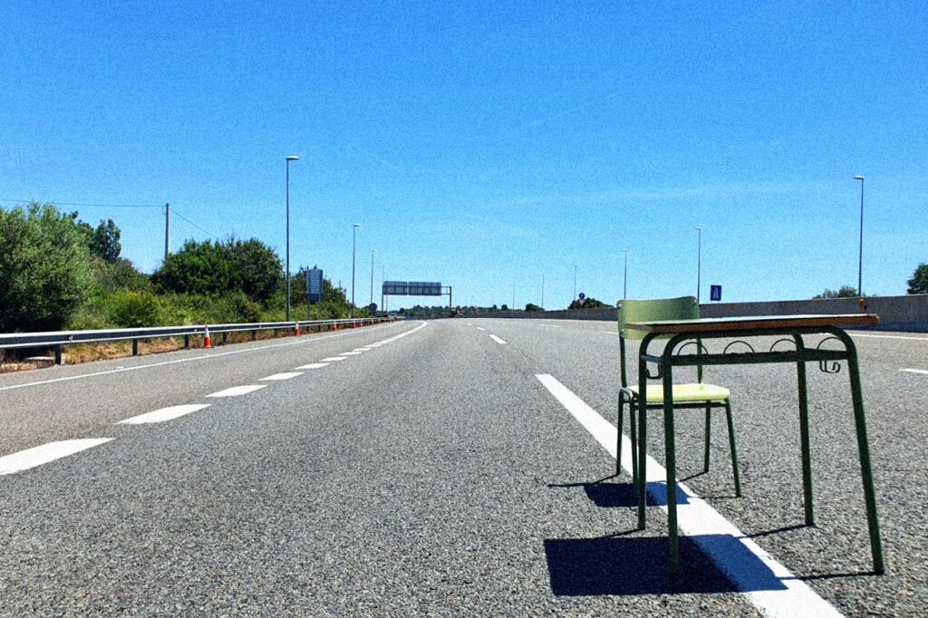 Taula i cadira de color verd d'una classe de centre educatiu, al mig d'una autopista buida.